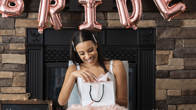 27 Bridal Shower Ideas That Feel Fresh, Fun, and Actually Personal 5 Smiling bride-to-be opens a gift bag at her bridal shower, with rose gold balloon letters spelling BRIDE in the background