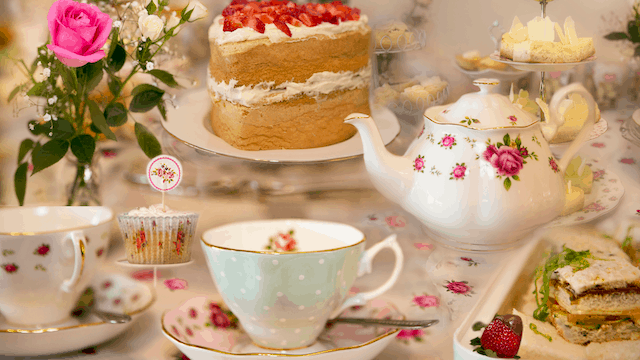 Elegant afternoon tea setup with floral china teapot, teacups, layered sponge cake with strawberries, cupcakes, and pastries on a decorated table.