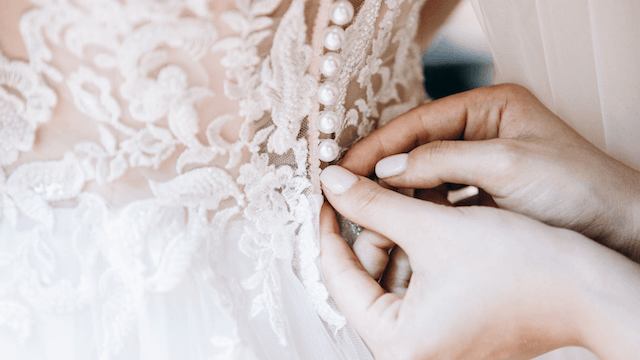 Close-up of hands buttoning the delicate lace back of a wedding dress with pearl details