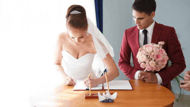 How To Plan A Wedding Without a Wedding Planner: 16 Tips 1 The bride in a white gown signs the wedding documents while the groom in a burgundy suit holds a bouquet of pink roses.