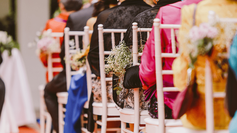 Wedding Advice 13 Guests are seated at a wedding ceremony, in colorful attire, and with floral arrangements.