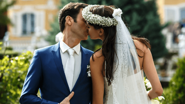 How to Deal With Parents Controlling Wedding Planning 4 Bride and groom sharing a kiss outdoors, with the bride wearing a lace veil and floral crown and the groom in a blue suit