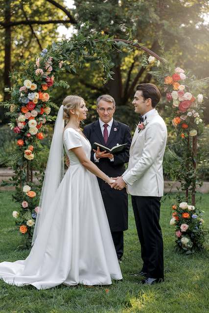 wedding officiant Bride and groom holding hands under a floral arch during outdoor wedding ceremony with officiant