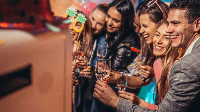 Group of wedding guests laughing and posing with props for a fun photo booth moment, holding champagne glasses.


