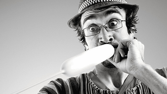 A man in glasses and a hat humorously inflates a long balloon, creating a playful, entertaining expression.
