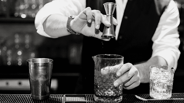 Wedding Vendor Meals: Feed Your Band & Photographer Without Going Crazy 4 Bartender pours liquid from a jigger into a mixing glass filled with ice beside a shaker and cocktail glass on the bar