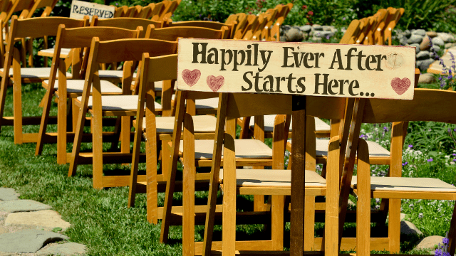 Post-Wedding Depression: When The Blues Set In after the Big Day 2 Outdoor wedding ceremony rows of wooden chairs with a rustic sign that reads “Happily Ever After Starts Here…” decorated with hearts.