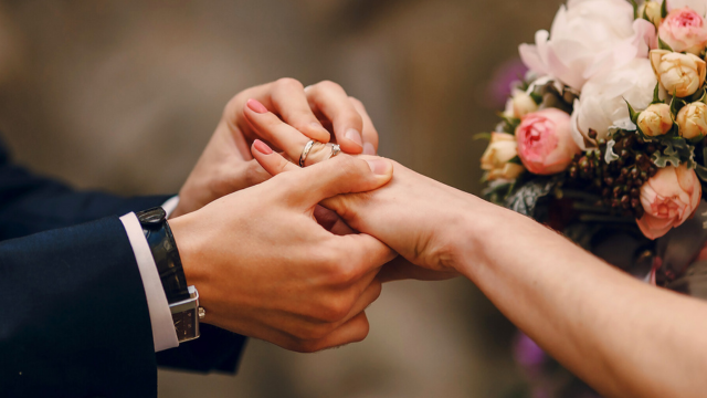 What Hand Does a Wedding Ring Go On? The Ultimate Guide 4 Groom slides wedding ring onto bride’s left ring finger beside a bouquet of pink peonies during the ceremony