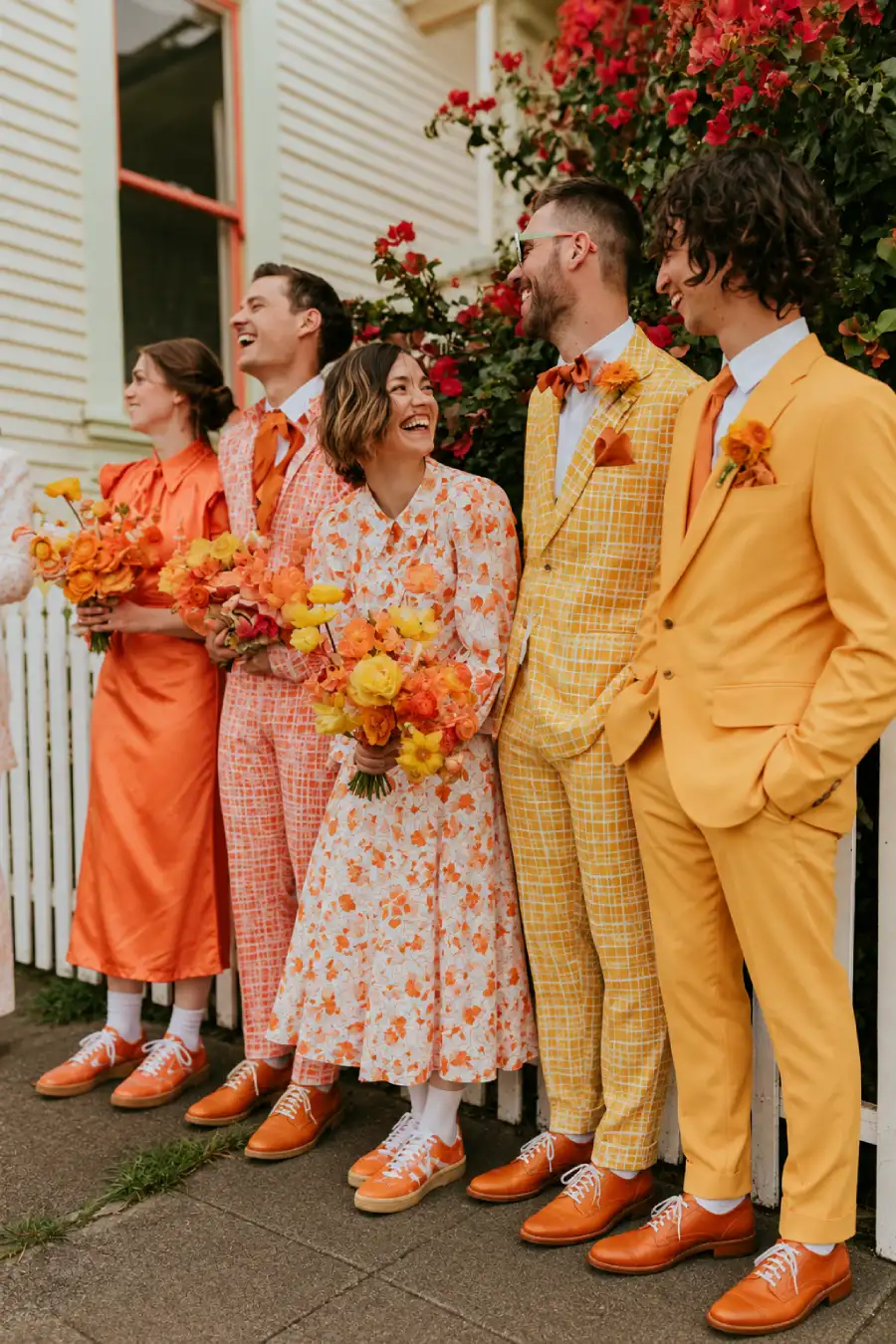 Groovy Wedding Theme: Creating a Retro Celebration 7 Wedding party in retro orange and yellow suits and floral dresses, holding bright bouquets outside a flower-covered house