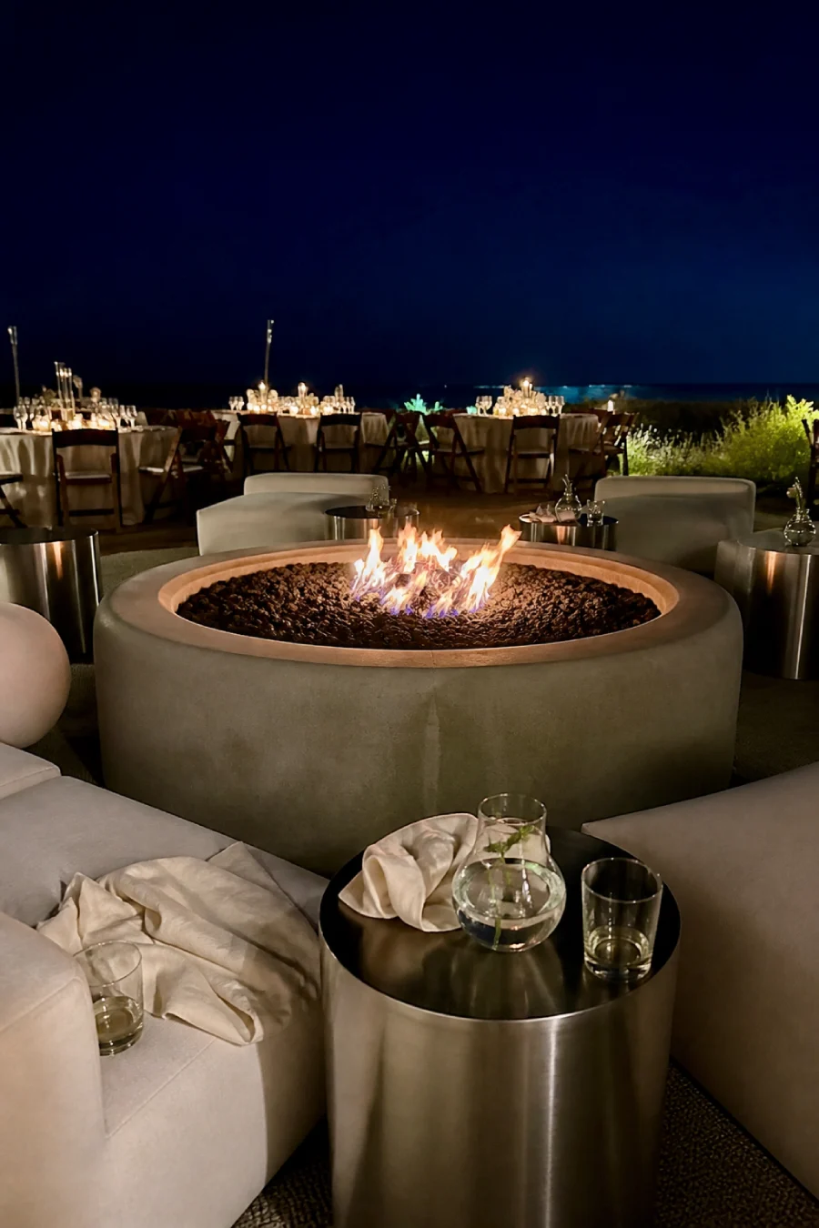 33 Jaw-Dropping Ways to Wow Your Wedding Guests at the Reception 9 Round firepit glows between ivory lounge seats and silver tables beside candlelit wedding tables overlooking the ocean below