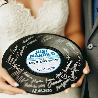 Bride and groom holding a signed vinyl record guest book that reads “Just Married” with wedding date and signatures, adding a retro music-themed keepsake touch
