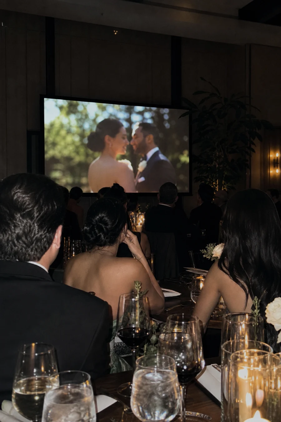 33 Jaw-Dropping Ways to Wow Your Wedding Guests at the Reception 10 Wedding guests watch the couple's love story video from candlelit dinner tables inside a dim contemporary reception ballroom.