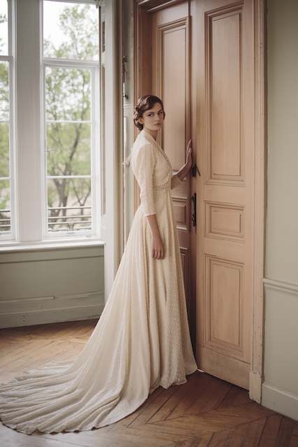 Bride in a vintage inspired long sleeve gown with delicate beading and flowing train stands by tall paneled doors in an elegant historic room with large windows and soft natural light