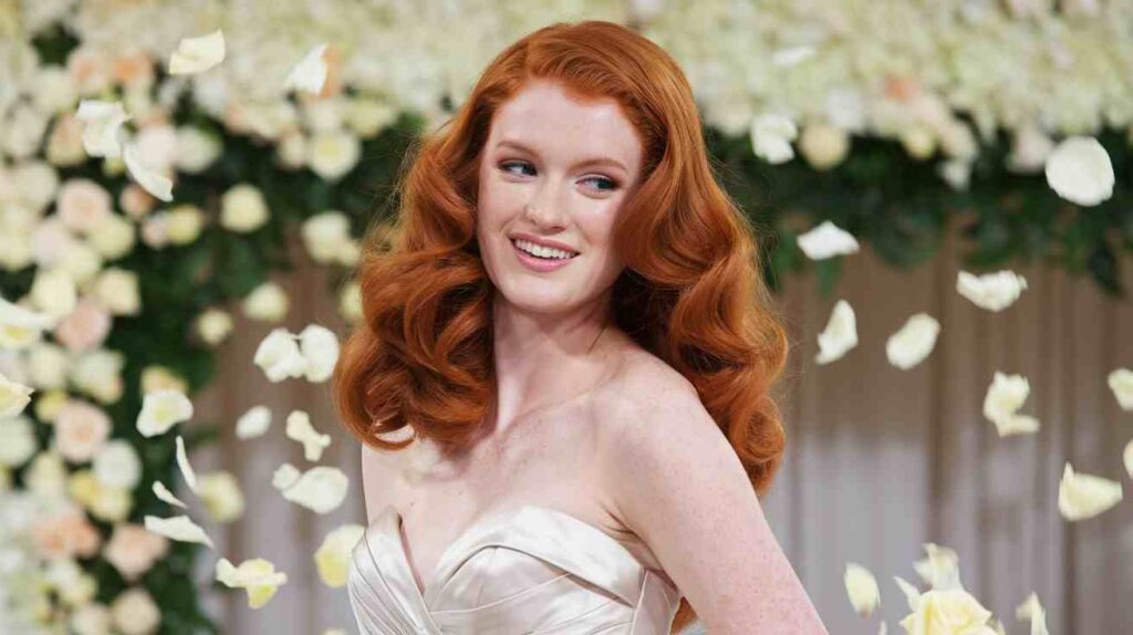 Face-Framing Soft Curls Bride with long red hair in Face-Framing Soft Curls, wearing a strapless ivory gown and smiling as rose petals fall, floral arch behind her