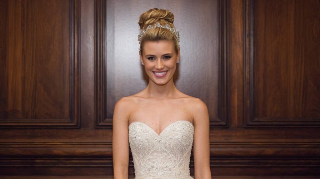 Top Knot with Crown Bride in strapless beaded gown wearing a high top knot with a crystal crown, standing before a wood paneled wall and smiling at the camera.