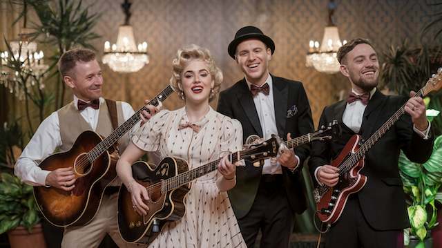 Vintage wedding band performing in retro attire, with female singer and musicians playing guitars in a classic ballroom setting with chandeliers and patterned wallpaper