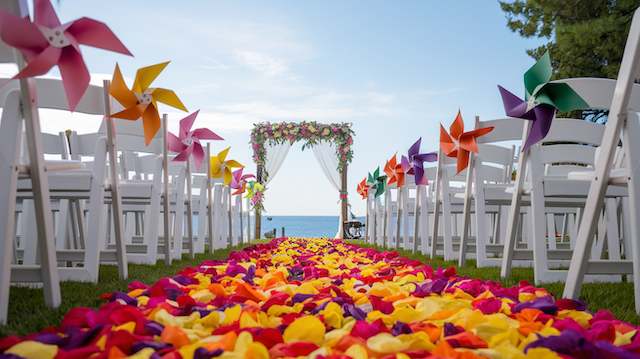 Paper Pinwheels Beachfront wedding aisle with white chairs, bright pinwheels, carpet of pink, orange, yellow petals, floral arch by the sea.
