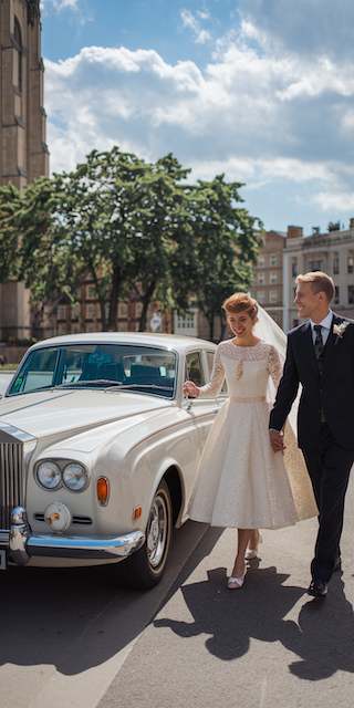 Elegant bride in a tea-length lace wedding dress and groom in classic black suit holding hands beside a vintage white Rolls-Royce on a sunny city street for a timeless retro wedding moment