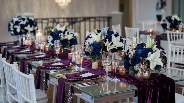 Rectangular mirrored tables with plum velvet runners, white Chiavari chairs, navy roses and white lilies centerpieces, glowing votive candles