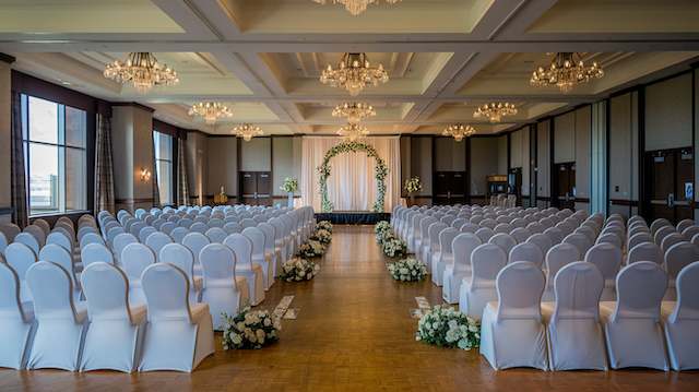 Banquet Chairs Banquet hall wedding setup with white banquet chairs covered and arranged in rows facing a floral arch under chandeliers.