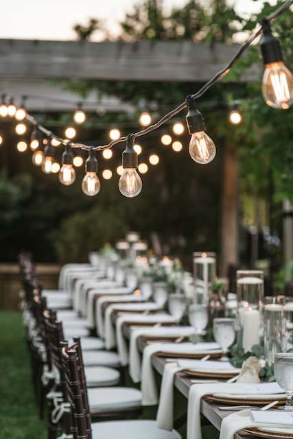 Bistro Style Patio Lights Warm bistro string lights hanging above a long outdoor wedding table set with candles, greenery, and elegant place settings at dusk.