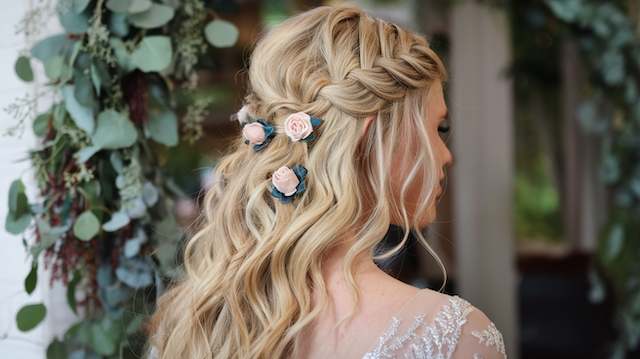 Cascading Side Braid Bridesmaid with a cascading side braid and loose curls, decorated with small blush flowers, standing beside greenery at an outdoor venue