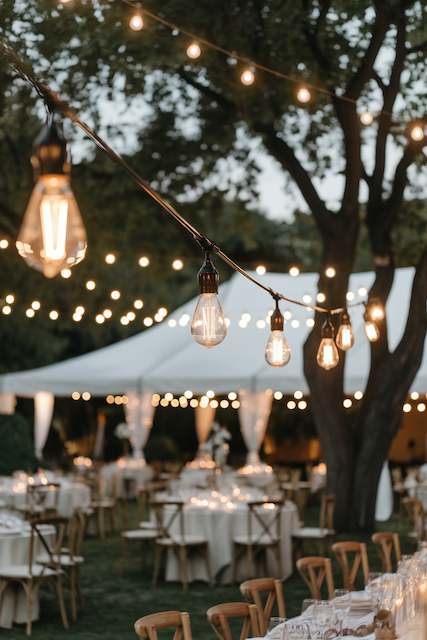 Edison Bulb String Lights Outdoor wedding setup with Edison bulb string lights hanging between trees, glowing warmly above a tented reception and dinner tables.