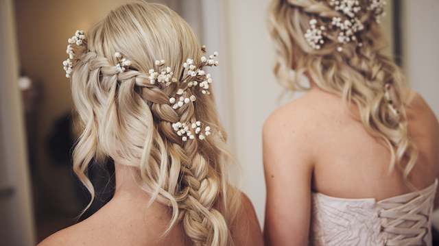 Floral-Accented Braids Two bridesmaids with blonde braided hairstyles decorated with clusters of baby’s breath, shown from behind in soft, natural indoor lighting.