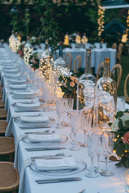 Glass Bottle Lights Long outdoor wedding table decorated with clear glass bottles filled with warm string lights, surrounded by flowers, candles, and elegant place settings.