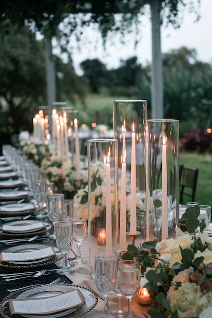 Hurricane Candle Holders Tall glass hurricane candle holders with glowing taper candles lining an elegant outdoor wedding table decorated with greenery and flowers.