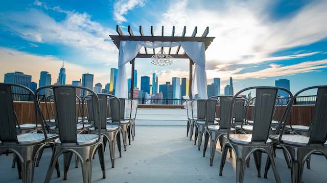 Industrial Metal Chairs Rooftop wedding setup with industrial metal chairs facing a fabric-draped pergola, city skyline visible in the background.