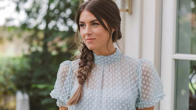 Loose Braided Ponytail Bridesmaid with a loose braided ponytail and soft face-framing strands wearing a light blue dotted blouse, standing outdoors by greenery.