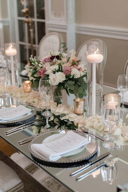 Mirror Tabletops Mirror tabletop with candles and a floral arrangement featuring roses and greenery, creating an elegant and reflective centerpiece.