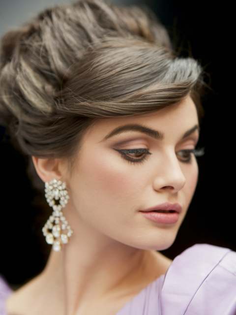 Retro Bouffant Bridesmaid with a voluminous retro bouffant updo and soft side-swept bangs wearing statement crystal earrings, shown in soft lighting.
