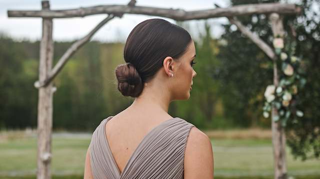 Sleek Low Bun Bridesmaid with a sleek low bun and deep side part wearing a taupe gown, standing outdoors beneath a wooden arch with greenery in the background