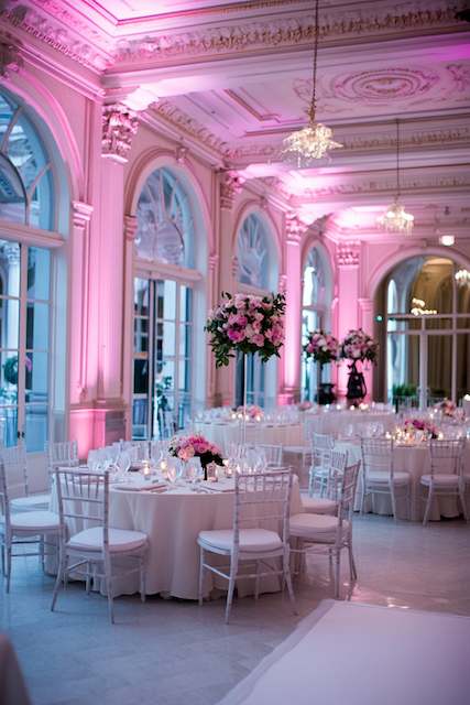 Soft Uplighting A luxurious ballroom with pink uplighting, white Chiavari chairs, and round tables adorned with tall floral centerpieces, creating an elegant wedding reception setting.
