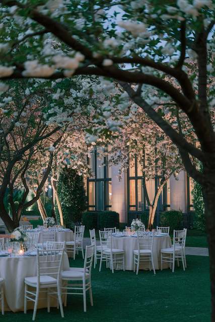 Spotlights on Trees Tree branches glowing with warm spotlight uplighting above elegant outdoor wedding tables with white chairs beside a modern glass venue.