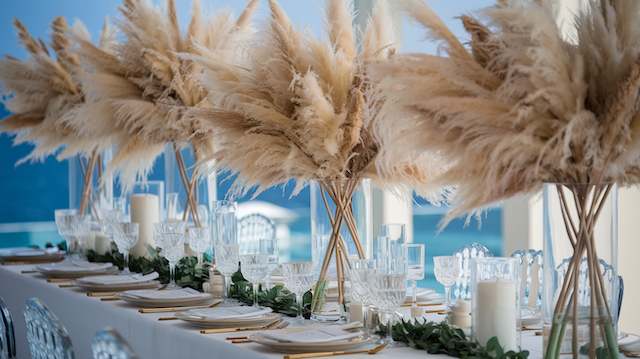 Tall Pampas Grass Arrangements Tall pampas grass centerpieces on a long wedding table with candles, glassware, and greenery, styled against bright windows overlooking the ocean.