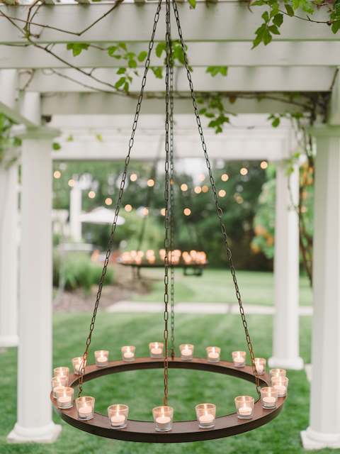 Tea Light Chandeliers Hanging tea light chandelier suspended from a pergola, creating a warm, romantic glow at an outdoor wedding reception with greenery in the background.