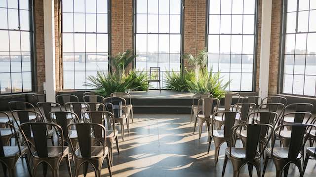 Tolix Chairs Industrial-style setup with metal Tolix chairs facing a stage with lush greenery and large windows showcasing a waterfront view.