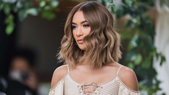 Wavy Lob Bridesmaid with a wavy lob styled in soft loose waves and a middle part, wearing an off shoulder cream top, standing outdoors near greenery.