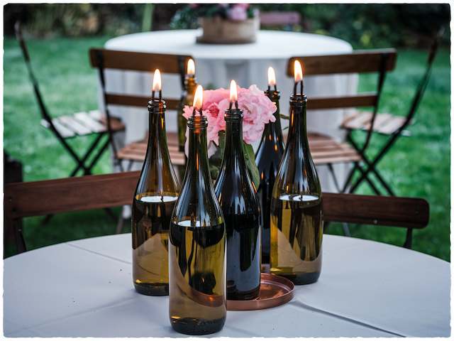 Wine Bottle Tiki Torches Wine bottle tiki torches with glowing flames arranged around pink flowers on an outdoor wedding table with wooden chairs in the background.