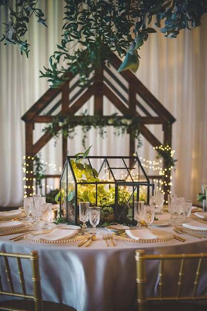 Mini glass greenhouse centerpiece filled with greenery and fairy lights on an elegant rustic wedding table with gold-rimmed plates and crystal glassware. Mini glass greenhouse centerpiece filled with greenery and fairy lights on an elegant rustic wedding table with gold-rimmed plates and crystal glassware.