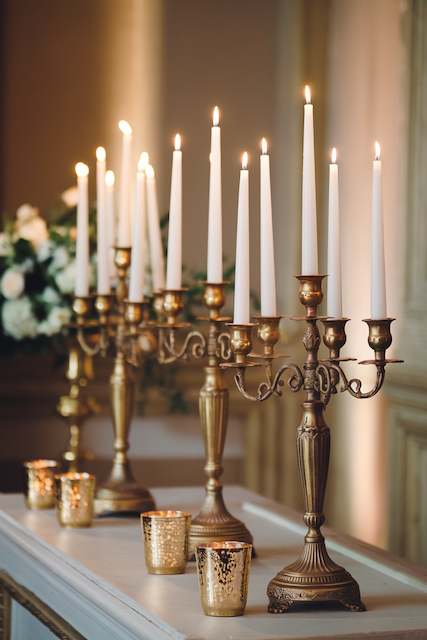Antique Gold Candle Stands For The Altar A line of ornate gold candelabras holding tall white taper candles creates an elegant and warm ambience. Smaller gold votive holders are placed in front, enhancing the sophisticated display.
