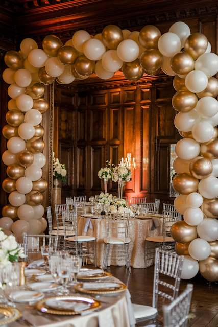 Balloon Garlands Balloon arch in gold and ivory creates a grand entrance to an elegant dining area with round tables, floral centerpieces, and Chiavari chairs.