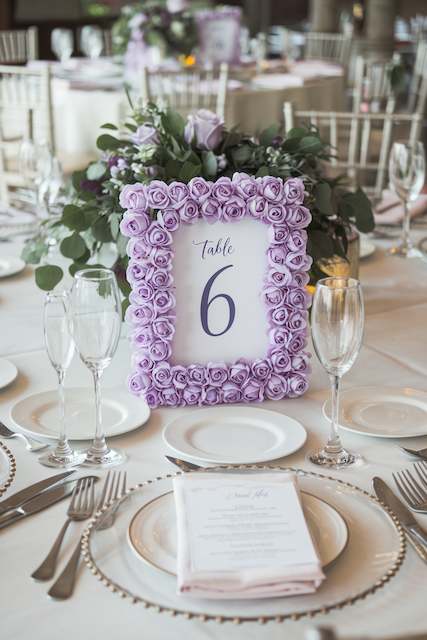 DIY Table Numbers A table setting with purple rose frame displaying "Table 6," surrounded by elegant dinnerware, glassware, and lush floral arrangements.