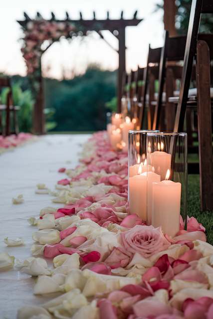 Flower Petals And Candles An outdoor wedding aisle lined with pink and white rose petals and glass cylinder candles, leading to a wooden pergola adorned with flowers.
