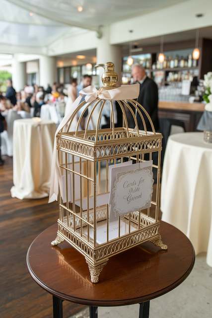 Gold Birdcage Card Box A gold birdcage serves as a card holder for wedding greetings. It sits on a wooden table, surrounded by guests in an elegant venue.