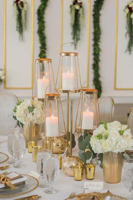 Gold Lanterns Clusters A sophisticated table setup featuring gold lanterns with white candles, surrounded by gold vases filled with white flowers. Elegant tableware and glassware complete the luxurious look.