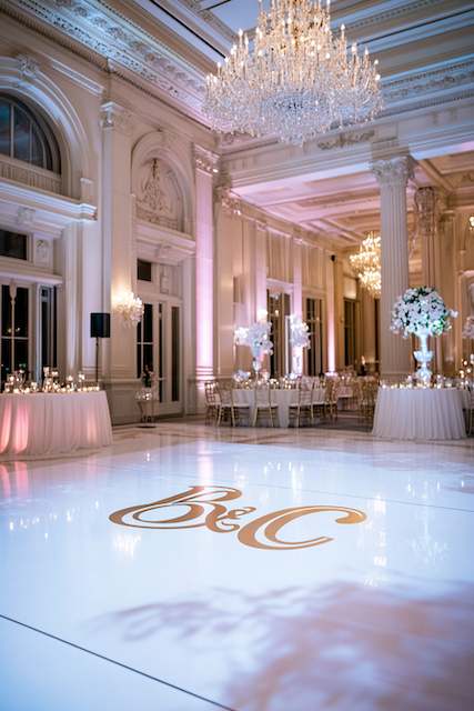 Gold Monogram Dance Floor Decal A grand ballroom with elegant chandeliers and tall windows features a monogrammed white dance floor. Tables with white and gold decor line the walls, enhancing the luxurious atmosphere.